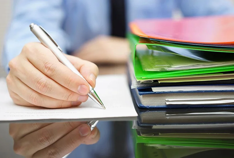 A person writing on paper with pens and papers.